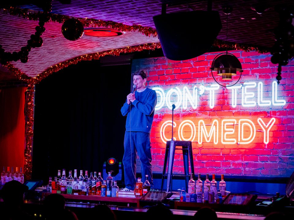 A comedian performs in front of a neon Don't Tell Comedy sign at On the Record inside Park MGM