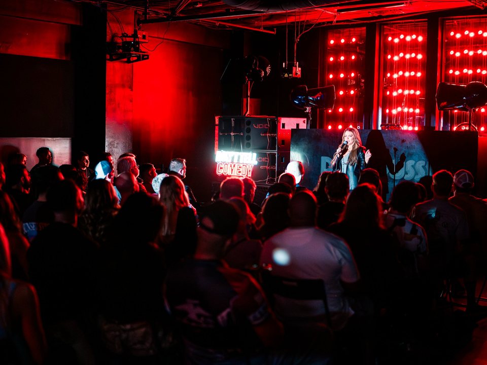 A comedian performs for a packed, red-lit crowd beside a neon Don't Tell Comedy sign at Discopussy