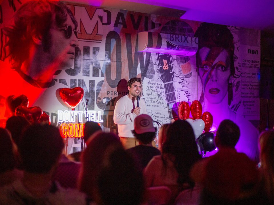 A comedian performs in front of a rock-poster mural and a neon Don't Tell Comedy sign at Fergusons Downtown